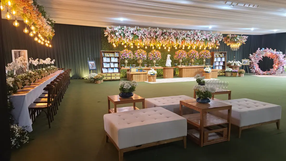 Salão montado para chá com mesa longa, poltronas e painel de flores